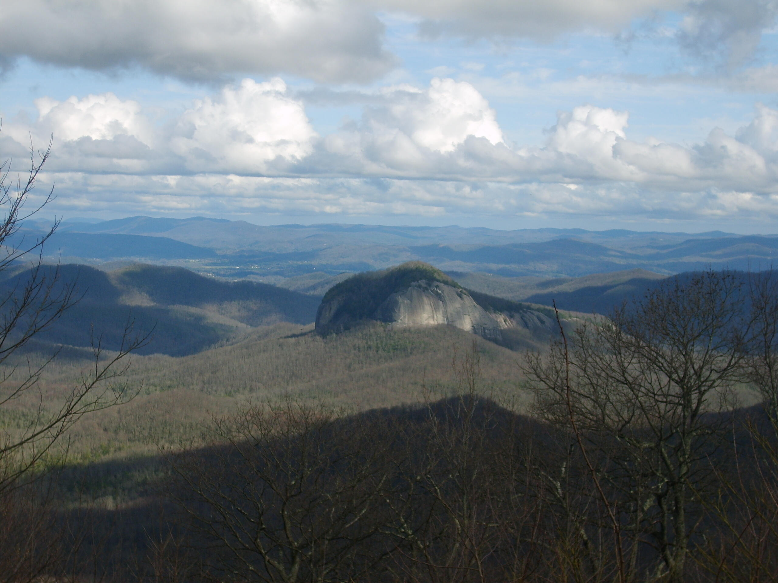 Rows of white cumulus clouds in a blue sky, over endless blue-green mountain peaks, with a prominent granite-faced, domed mountain in the middle, and leafless trees arching into the frame in the foreground